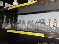 Wide view of 13 assorted crystal and decorative bells arranged on a dark wood shelf.