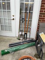 Photo showing a hole digger, spade, two other hand tools with wooden handles, and three rolls of netting (1 green deer netting and 2 black plastic netting rolls).