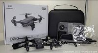 Deerc D20 drone in front of its box, remote controller, carrying case, spare blades in plastic bag, and charging cables arranged on a white surface.