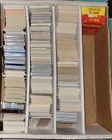 Photo of a box divided into three compartments containing numerous stacks of baseball cards, some sealed in plastic and some loose, including a red labeled box from 1990 Series 1 visible in right compartment.
