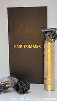Photo shows a gold metal hair trimmer with a professional T blade head next to its black box labeled 'HAIR TRIMMER' and a USB charging cable wrapped in plastic.