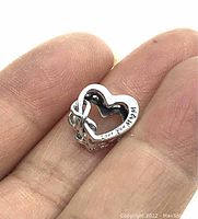 Close-up image of the sterling silver heart shaped bead charm held between fingers, showing the 'Love You Mum' engraving and intricate heart cutout details.