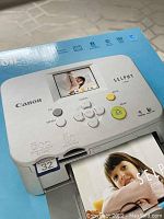 Close-up of Canon Selphy CP760 photo printer on retail box showing top controls and small LCD screen.