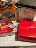 Photo showing boxed MiniDVD drive, boxed Verbatim Portable Hard Drive 160GB, boxed Harman Kardon Drive+Play, and boxed red Slingbox device.