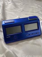 Blue Saitek digital chess clock with two LCD screens and control buttons on fabric background.