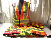 Front view of multiple neon orange safety vests with reflective yellow and silver stripes, folded showing role markings including Coordinator, Area Coordinator, and Captain.