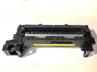 Front angle view of black plastic fuser assembly component for HP M607 LaserJet printer showing gear section and warning sticker.