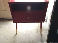 Front view of red wooden side table with drawer and open compartment showing wear on wood and hole for cords