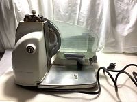 Side view showing stainless steel blade, food carriage with black handle, and white base