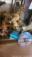 Photo showing multiple carousel horse figurines including a brown horse with blue hooves, cream-colored rearing horse, and a large grey carousel horse in the background, along with a blue trinket box and round decorative plate.