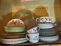 Stacked plates, cups, and covered bowl with botanical strawberry and butterfly design on white earthenware. Pale green plates behind.