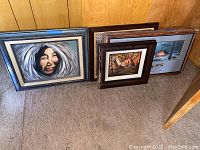 Four framed art pieces leaning against a wood-paneled wall showing diverse styles including portrait and landscape paintings.