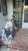 Photo displaying multiple garden statues placed on a brick floor including dog statues, a cowboy statue, boots with skulls statue, and a rabbit statue, along with a vertical butterfly and leaf metal hanging on the door.