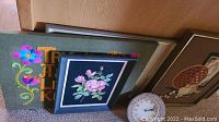 View of the two needlepoint wall art pieces stacked and the clock with pearl decoration on the floor. The needlepoint art includes a pink rose design and colorful floral text design.