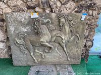 Front view of large rectangular plaster wall sculpture showing two raised horses with flowing manes, displayed against a stone wall background.