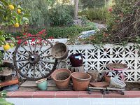 Wide view showing vintage wooden wagon wheel leaning against garden wall with assorted garden pots arranged nearby. Various materials and sizes of pots visible. Some pots are clay or ceramic. Metal pot mover visible.