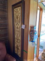 Wall hanging vertically framed with detailed floral and vine symmetrical design in brown and cream tones against a wooden panel background.