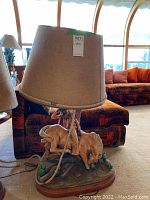 Front and side angle showing lamp base with cowboy and bull figurine under beige fabric lampshade with rope trim.