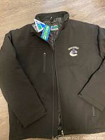 Front view of black Vancouver Canucks jacket with zipper, logo visible on left chest, tags attached.