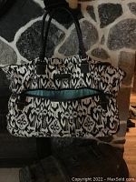 Front view of cloth duffle bag with black and white abstract pattern and black handles, turquoise interior visible in exterior zippered pocket.