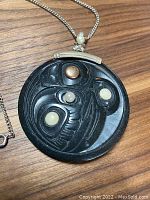 Front view of carved round black argillite pendant with gold and pearl inlays on silver chain