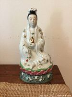 Front view showing seated Buddha figure in white hand painted porcelain with bamboo leaf details, holding a golden bottle and prayer beads, atop lotus and plinth base.