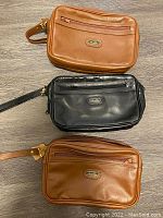 Three genuine leather bags (two tan, one black) arranged on floor showing front side with zipper pockets and wrist straps