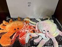 Five TY plush toys displayed on black mesh surface: three orange and pink Wiggly Beanie Babies and two Beanie Buddies in pastel colors and orange, all with TY heart tags.