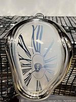 Front angled view of a melting style metallic silver wall clock, distorted face with Roman numerals.