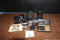 Full view of all vintage camera items on wooden surface showing Nikon camera body, Minolta lens, two light meters, Minolta electronic flash, lens cap, and instruction manual.