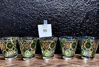 Front view of six glass tumblers arranged side by side showing the green and gold floral pattern on the clear glass.