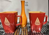 Two square vintage coffee mugs in burnt orange with cream interior and white geometric design, alongside a bright orange slender vase.