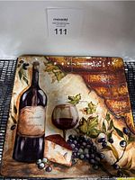 Square ceramic serving dish with a painted design featuring a bottle of Cabernet Sauvignon, a wine glass with red wine, grapes, and grape leaves against a rustic brick background.
