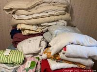 Stack of assorted bed linens including blankets, sheets, and towels in various colors and textures piled on a surface.