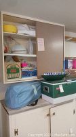 Photo showing three shelves filled with camping goods including a vintage Coleman cooler below the cabinet, a green Coleman drink cooler, white plates, and a blue Aerobed bag.