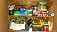 Wide view of shelf showing a Coleman propane fuel can, a blue lantern, a camouflage fishing gear bag, a glass jar with fishing hooks, various boxes and supplies.
