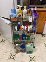 Full front view of wire 3 level cart filled with various cleaning spray bottles, liquid soap bottles, and other supplies on a kitchen floor.