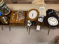 Overall view of assorted wall and desk clocks arranged on two tables, showing a variety of designs including wooden wall clock, small brass desk clock, electronic alarm clock, and clear plastic gear clock.