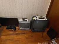 Wide shot showing three electronics items: one flat screen monitor on the left, small silver CRT TV in the middle, larger black and gray Zenith VHS TV on the right, all set on wooden floor