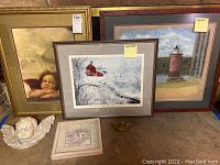 Framed cherub painting, framed winter barn scene, franes lighthouse seascape, and two 3D angel decorations on the floor in front.
