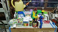 Photo displaying a variety of garden supplies including watering can, seed packets, liquid nutrient bottles, and fertilizer packages arranged on a wooden surface.
