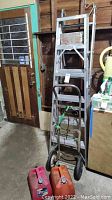Full view showing 6-foot aluminum ladder, metal hand truck with green strap, and gas cans nearby in rustic garage setting.