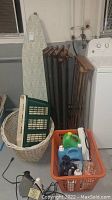Laundry essentials grouped together showing basket, ironing board, drying racks, and detergents near a washing machine.