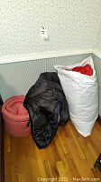 Three sleeping bags on a hardwood floor, one black bag laying loose, a red/brown rolled bag and a white storage sack with red interior visible.