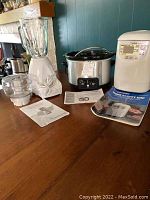 The lot shows three small kitchen appliances including a blender with glass jar and plastic food processor attachments, a stainless steel slow cooker with glass lid, and a white bread maker with control panel and manuals.