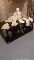 Full view of vintage trunk with cloth doll seated on top surrounded by pairs of ladies gloves
