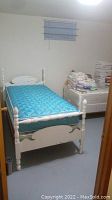 Painted white wooden twin bed assembled with blue mattress in a room with white walls and gray floor. Shows headboard, footboard and part of frame.