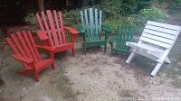 Group photo showing two red Adirondack style chairs, two green Adirondack style chairs, and one white slatted wooden chair arranged outdoors on a dirt and stone surface.