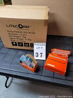 Box of 12 LiteCorr PAR38 LED bulbs, 15 orange boxes of J 130V 100W tungsten halogen lamps, and 5 smaller boxes of blue-white packaged T3 100W bulbs arranged on a table.