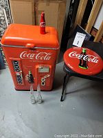 Overview of Coca-Cola plastic cooler with metal button sign on chair and bottles in front.